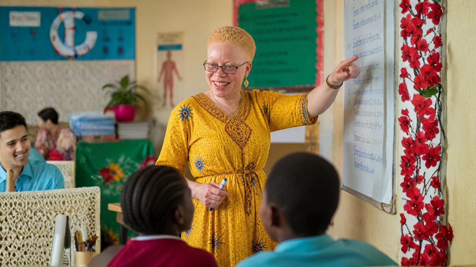 A teacher with albinism pointing at a whiteboard while teaching a Tanzanian classroom